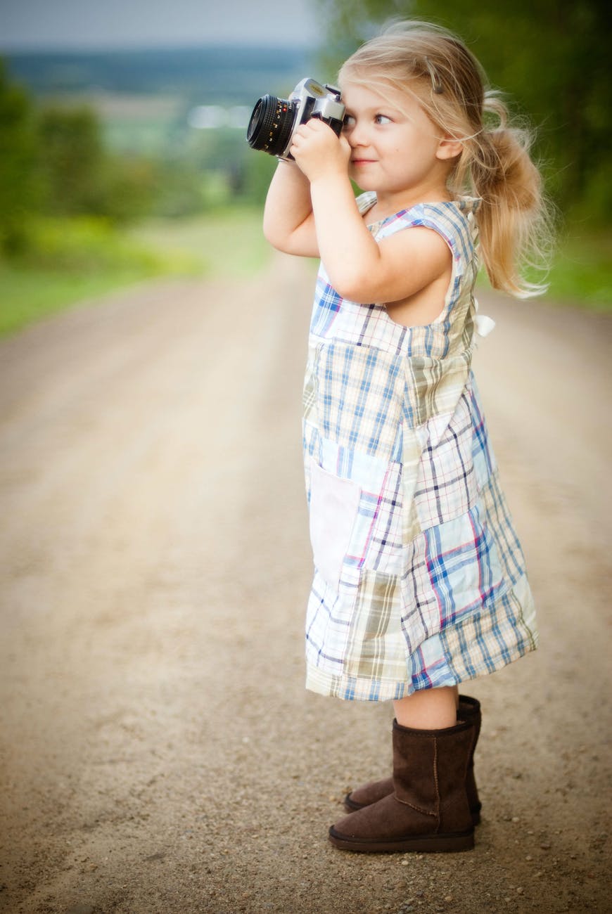 girl with blonde hair and wearing blue and white plaid dress and capturing picture during daytime